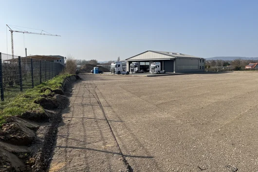 Baufortschritt beim Neubau eines Speditionsbetriebes in Hüllhorst mit befestigter Logistikfläche, Zaunanlage und Hallengebäude im Hintergrund